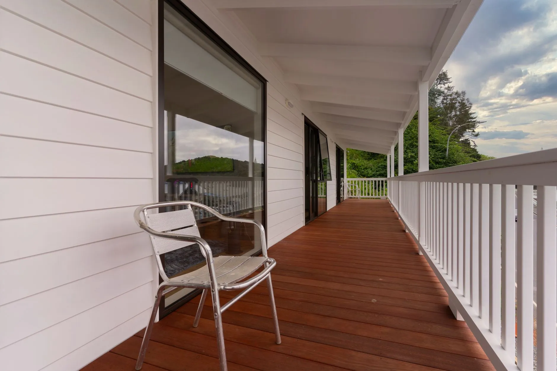 Covered porch deck with warm-toned wood boards, white railings and a metal chair